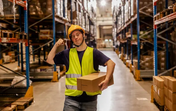 Down sendromlu kendine güvenen bir depo çalışanı dolu bir depoda kutu taşırken kolunu esnetiyor..