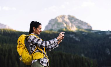 Sarı sırt çantalı gülümseyen kadın, dağ manzaralı bir bölgede yürürken telefonuyla fotoğraf çekiyor..