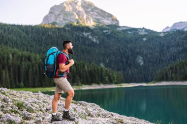 Mavi sırt çantalı bir adam orman ve kayalık araziyle çevrili bir dağ gölü manzarasına hayran olmak için duruyor..