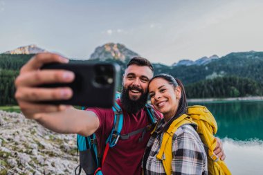 Sırt çantalarıyla donatılmış mutlu bir çift yürüyüş maceraları sırasında manzaralı bir dağ gölünün kenarında selfie çeker..