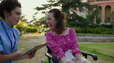 Bakıcı, parktaki açık hava etkileşimi sırasında elinde bir pano olan tekerlekli sandalyedeki genç bir kadınla konuşuyor..