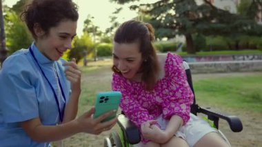 Bakıcı, açık havada güneşli bir anda tekerlekli sandalyede gülümseyen genç bir kadınla akıllı telefonunda bir şey paylaşır..