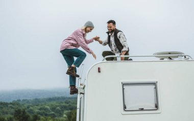 Two friends helping each other climb onto the roof of an RV, enjoying a fun moment during a camping trip. clipart