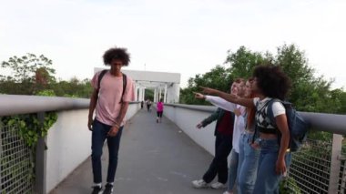 Bir grup öğrenci, köprüde tek başına yürüyen bir öğrenciyi işaret ediyor ve gülüyor. Alay ve zorbalık sahneleri yaratıyor..
