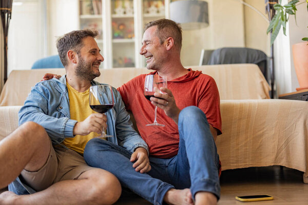 Улыбающаяся пара, сидящая на полу, разделяя тост с красным вином, наслаждаясь расслабленным моментом вместе дома.