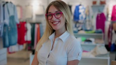Pembe gözlüklü gülümseyen genç satıcı moda bir giyim mağazasında güvenle duruyor..