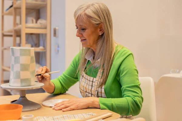 Акцентированная пожилая женщина тщательно рисует клетчатый дизайн на керамической вазе в художественной студии.