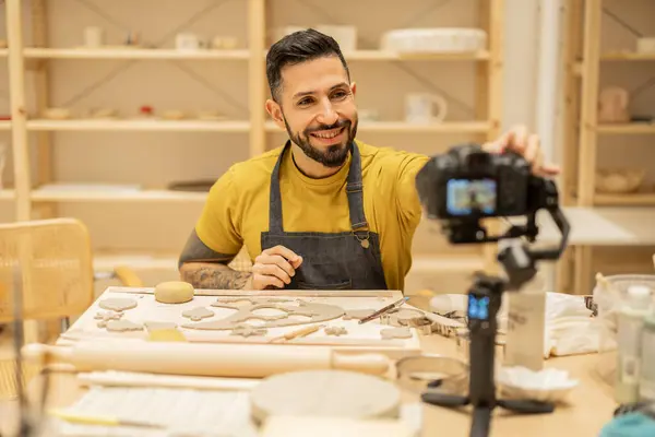 stock image Smiling man filming a pottery tutorial in a studio while shaping clay with tools on the table.