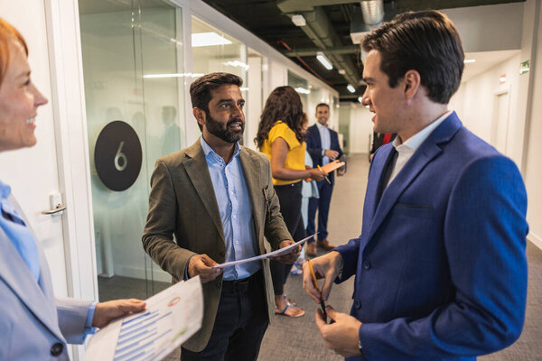 Группа профессионалов мультирасового бизнеса беседует в офисном коридоре, обменивается документами.