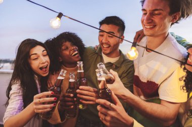 Bir grup genç yetişkin birayla kadeh kaldırıyor, tel ışıklarının altında eğlenceli bir çatı partisinin tadını çıkarıyorlar..