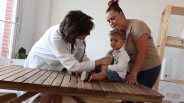 Bayan çocuk doktoru, çocuğun sağlığı hakkında annesiyle tartışırken, çocuk masada bir oyuncakla oynuyor..