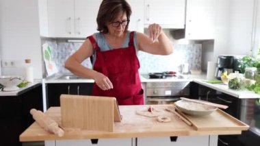 Kırmızı önlüklü bir kadın, parlak bir mutfakta hamurun üzerine un serpiyor. Ev yapımı fırınlanmış malların tarifini takip ediyor..