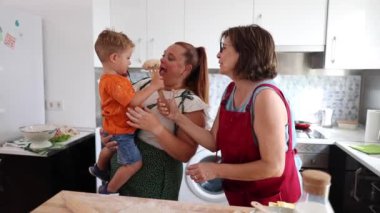 Anne ve büyükanne, mutfakta kaşıkta hamur tutan bir çocukla eğlenceli bir yemek pişirme anını paylaşıyor..