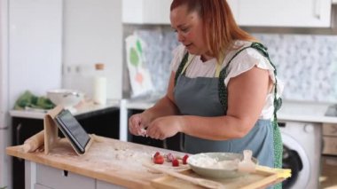 Kadınlar yemek pişirme ve teknolojiyi dengelemek için bir evde hamur ve çilek hazırlarken, yemek tarifi için tablet kullanırlar..