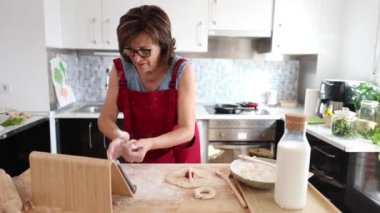 Kırmızı önlüklü kadın mutfaktaki bir tabletten yemek tarifi alırken hamur yoğuruyor, ev yapımı pişirilmiş ürünler hazırlıyor..