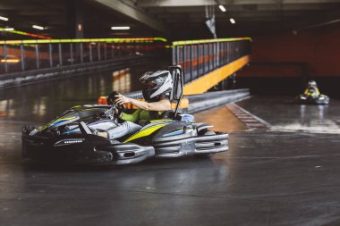 Kasklı genç bir kadın kapalı pistte go-kart kullanıyor, arka planda başka bir kartla yarışıyor..