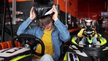 Kot gömlekli bir çocuk kaskını giyiyor ve kapalı bir pistte go-kart yarışına hazır. Genç Çocuk Go-Kart Yarışı için Miğfer Takarak Hazırlanıyor