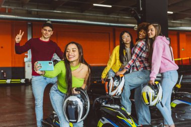 Bir grup arkadaş, ellerinde kask olan ve kapalı bir go-kart pistinde gülümseyen bir selfie için poz veriyorlar..