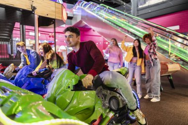 Group of friends enjoying an arcade motorcycle racing game, with others cheering and taking photos in the background. clipart