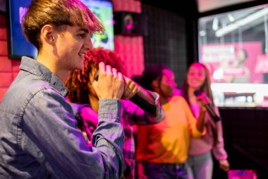 Bir grup arkadaş canlı bir odada karaoke söylüyor, gülümsüyor ve mikrofon tutarken eğleniyorlar..