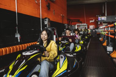 Bir grup genç yetişkin, kapalı bir yarış pistinde go-kart sürüyor, gülümsüyor ve eğleniyorlar..