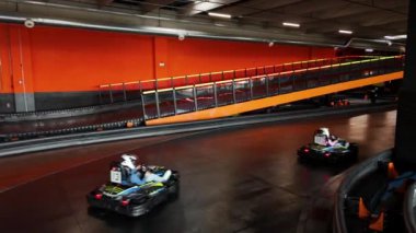 İki go-kart, canlı turuncu arka planı ve rampası olan kapalı bir pistte hız yapıyor..