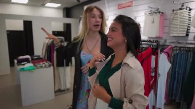 Moda mağazasında alışveriş yapan iki mutlu genç kadın. 20 'li yaşlarda iki genç kadın bir butikte neşe içinde kıyafetlerini karıştırıyor, kahkahalarını paylaşıyorlar..