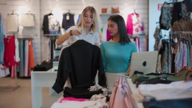 İki genç kadın birlikte alışveriş yapıyor, ellerinde siyah bir kıyafet, ve bir butikte moda tarzlarını tartışıyorlar. İki genç bayan alışveriş asistanı ve müşteri Kara Cuma günü bir giyim mağazasında moda seçeneklerini tartışıyor.