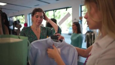 Kadın, moda mağazasından kıyafet alırken akıllı telefonuyla ödeme yapmaya hazırlanırken gülümsüyor. Kara Cuma günü bir giyim mağazasında satış elemanı ve müşteri