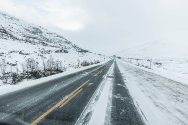 Karla kaplı boş bir yol, kışın karla ve engebeli araziyle çevrili uzak dağlık bir arazide uzanır..
