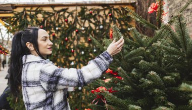 Bayram süsleriyle çevrili, açık hava pazarında Noel ağacı seçen bir kadın..