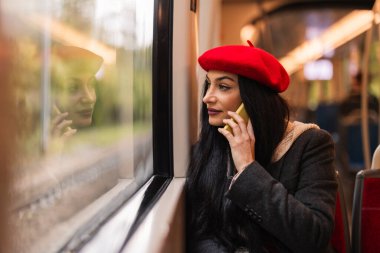 Kırmızı bereli bir kadın, tren penceresinin yanında oturup dışarıyı seyrederken telefonuyla konuşuyor..