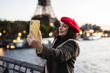 Smiling woman in a red beret taking a selfie with her phone near the Eiffel Tower at dusk, capturing the illuminated Parisian skyline. clipart