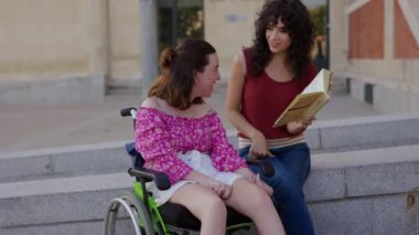 Tekerlekli sandalyede beyin felci geçiren bir kadın arkadaşıyla bir kitabı paylaşırken gülümsüyor, açık hava bölgesinde birlikte oturuyor..