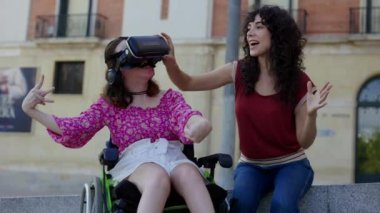 Tekerlekli sandalyede beyin felci geçiren bir kadın, arkadaşının da desteğiyle şevkle VR kulaklık kullanıyor..