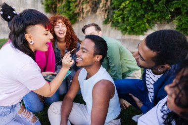 A diverse group of friends enjoys applying makeup on a smiling non-binary man during an outdoor gathering. clipart