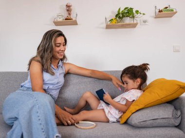 Gülümseyen anne, kanepede birlikte dinlenirken küçük kızının akıllı telefonuyla meşgul oluşunu izliyor. Anne ve kızı akıllı bir telefonla kanepede dinleniyorlar.