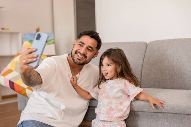 Gülümseyen baba ve kız, eğlenceli bir selfie için poz veriyor, oturma odalarında neşeli ve neşeli bir anı yakalıyorlar..