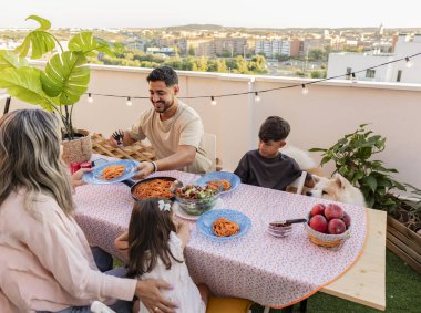 Gülümseyen aile balkonlarında spagetti ve salata yer, günbatımının tadını çıkarır ve birlikte vakit geçirirler..