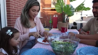 Latin Amerikalı bir aile, güneşli bir günde terasta oturmuş taze bir salatayı paylaşıyorlar.