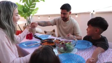 Gülümseyen Latin Amerikalı aile, köpekleri yakınlarda otururken neşe içinde el ele tutuşup avluda yemek yiyorlar..