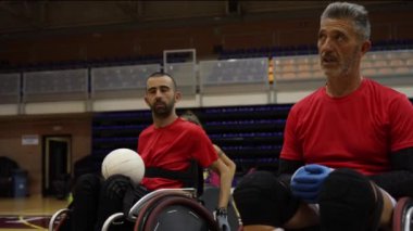 Kırmızı forma giyen iki tekerlekli sandalyeli rugby oyuncusu, kapalı alanda antrenman yaparken topu tutuyorlar..