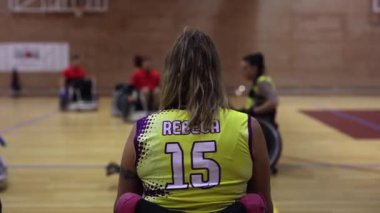 Basketbol sahasında tekerlekli sandalyesinde oturan sarı ve mor üniformalı bir sporcu maçın başlamasını bekliyor.