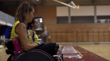 Engelli sporcular oynamak için sıralarını bekliyorlar. Basketbol sahasının kenarındaki tekerlekli sandalyelerde oturuyorlar.