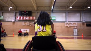 Tekerlekli sandalyede oturan bayan basketbolcu, takım arkadaşlarının antrenmanlarını izliyor.