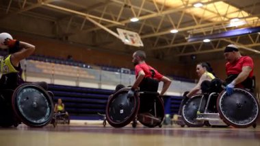 Tekerlekli sandalyedeki felçli atletler, parlak ışıklandırılmış bir spor salonunda, takımla strateji oluşturuyorlar.