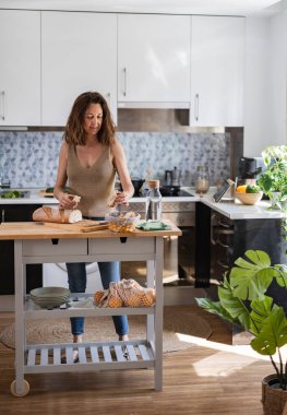 Kadın modern mutfağında duruyor, ekmek ve meyveli sağlıklı bir kahvaltı hazırlıyor. Parlak alan doğal ışıkla dolu.