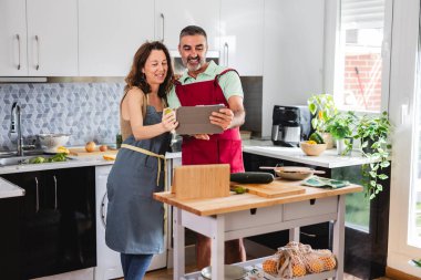 Önlük giyen olgun bir çift, mutfakta birlikte yemek tarifi hazırlar, tablet üzerinde talimatları izler ve iyi vakit geçirirler.