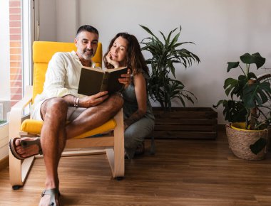 Erkek ve kadın, rahat oturma odalarında doğal ışık ve bitkilerle dolu bir odada huzurlu ve sevgi dolu bir atmosfer yaratarak birlikte okumanın keyfini çıkarırlar.