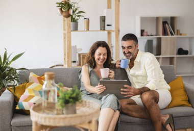 Gülümseyen çift evlerinde rahat bir sabahın tadını çıkarırken, rahat koltuklarında oturup tabletlerinde interneti karıştırıyor ve kahvelerini yudumluyorlar.